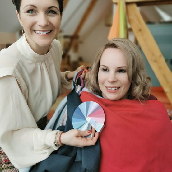 Zwei lächelnde Frauen halten ein Farbrad und stehen in einem Raum mit Holzwänden. Eine Frau hält einen roten Stoff.