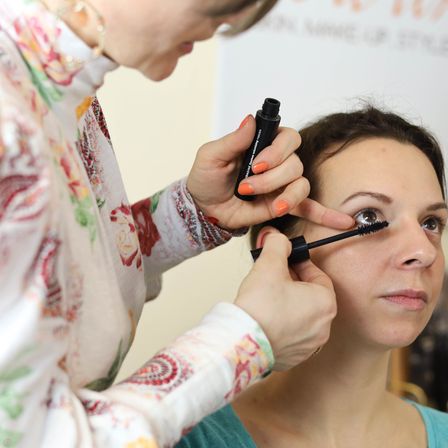 Eine Frau trägt einer anderen Frau Mascara auf die Wimpern auf. Die Frau, die Mascara aufträgt, trägt ein Blumenhemd.