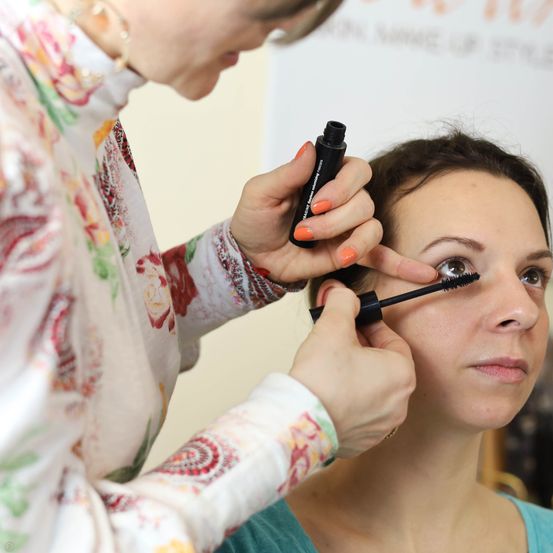 Eine Frau trägt einer anderen Frau Mascara auf die Wimpern auf. Die Frau, die Mascara aufträgt, trägt ein Blumenhemd.