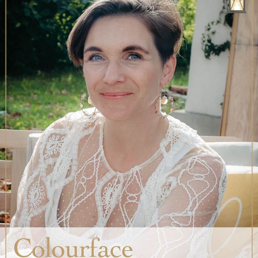 Eine Frau mit kurzen Haaren und blauen Augen lächelt und trägt ein weißes Spitzenoberteil und Perlenohrringe. Der Hintergrund zeigt einen Rasen mit Pflanzen.