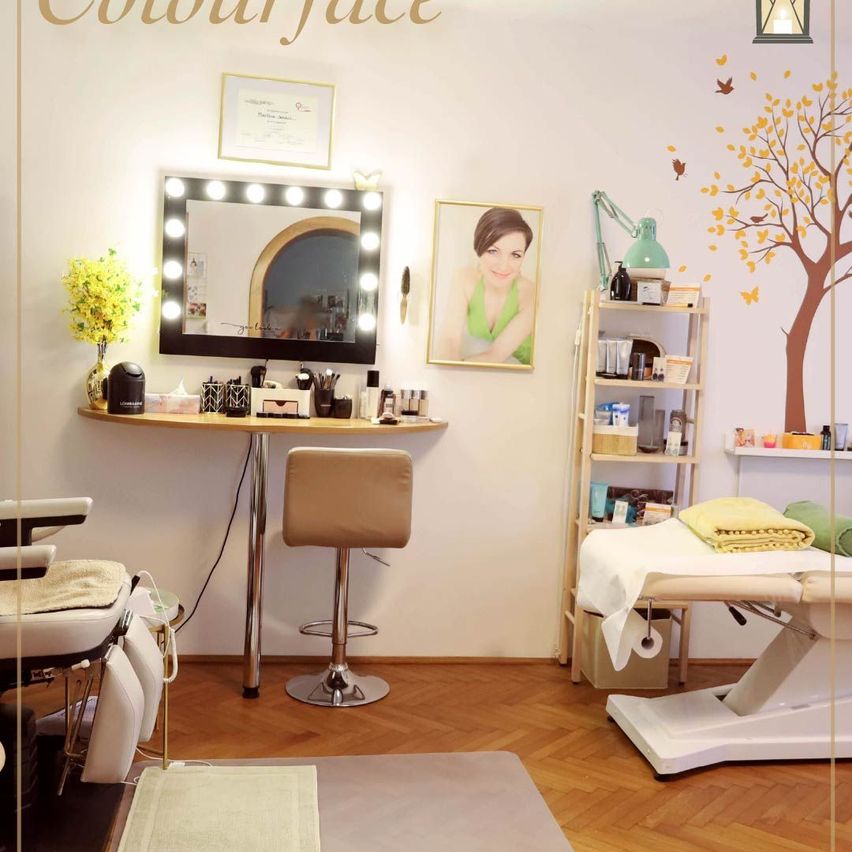 Innenraum eines Schönheitssalons mit einem Schminkspiegel mit Lichtern, einem Hocker und einem Bett rechts. An der Wand befinden sich ein Bilderrahmen, ein Baumdesign und eine Lampe. Der Boden ist aus Holz und es gibt eine gelbe Blume in einer Vase.