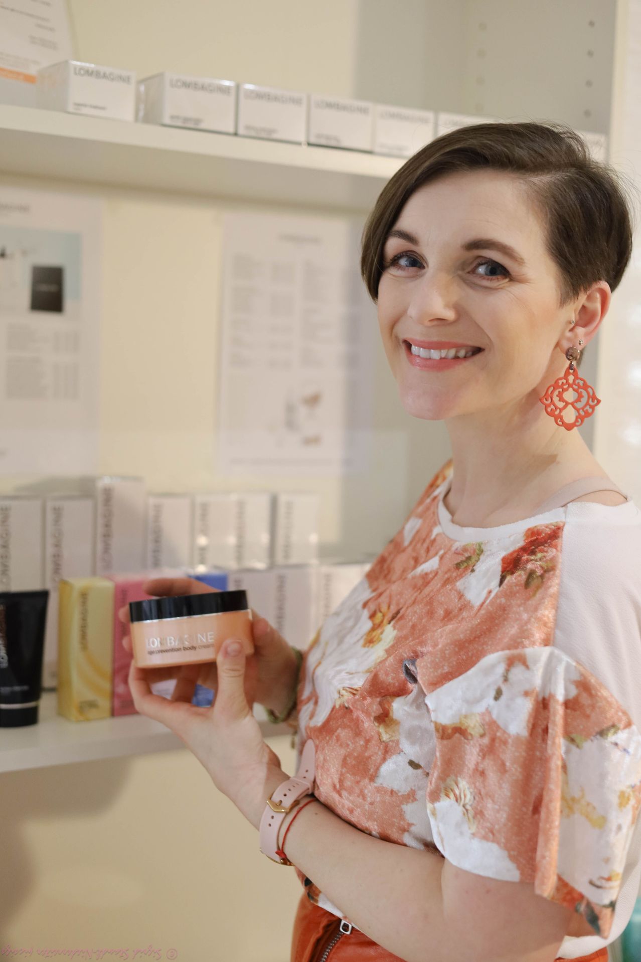 Eine lächelnde Frau hält eine Dose Hautcreme und steht vor einem Regal mit verschiedenen Produkten in einem gut beleuchteten Raum.