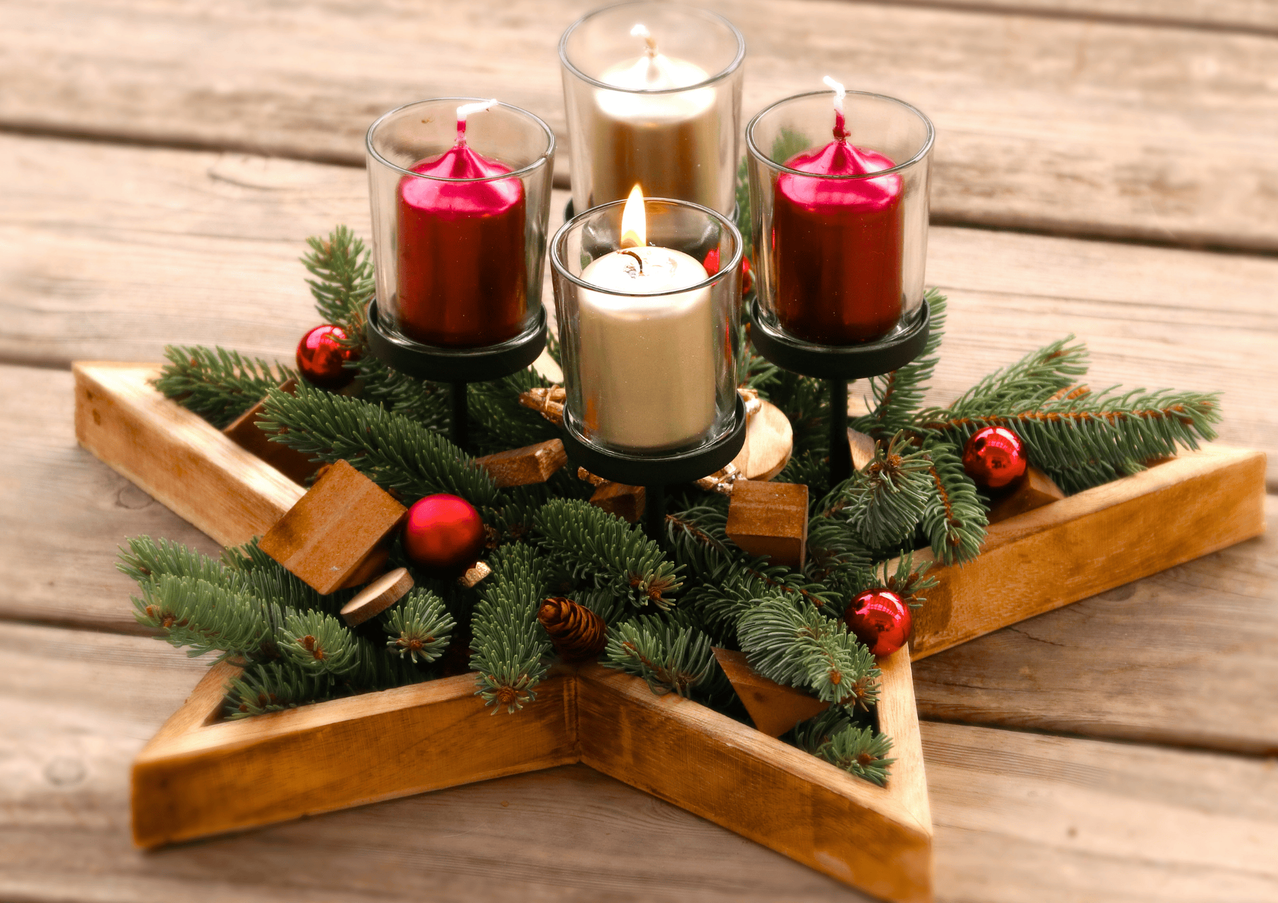 Eine sternförmige Holzdekoration hält fünf Kerzen, zwei rot und drei weiß, umgeben von immergrünen Zweigen und roten Kugeln auf einer Holzoberfläche.