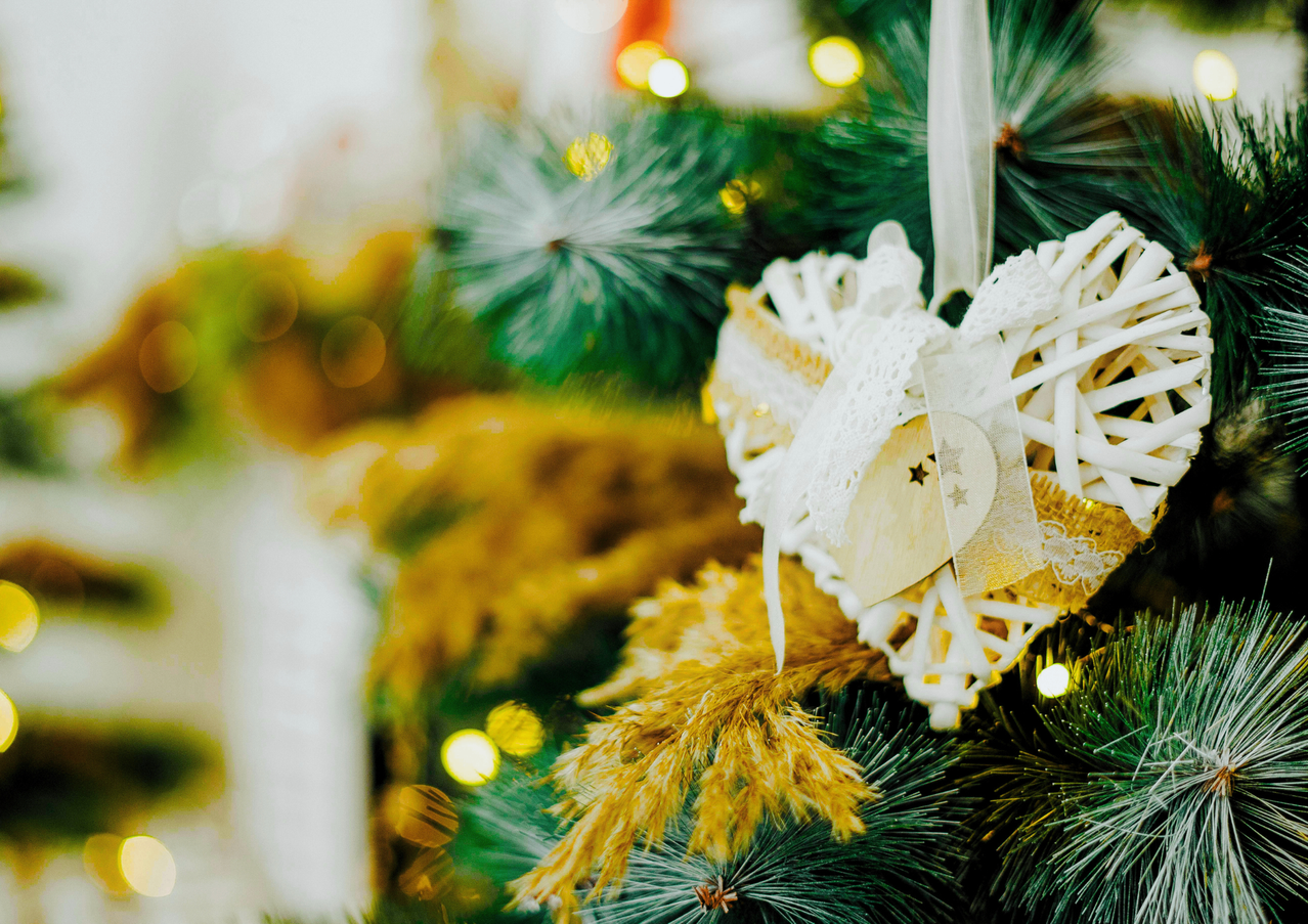 Ein Nahaufnahme einer Weihnachtsbaum, geschmückt mit einem herzförmigen Ornament. Das Ornament ist aus weißer Spitze und einem kleinen Holzkreis gefertigt. Der Baum hat gelbe und grüne Blätter und einige funkelnde Lichter.