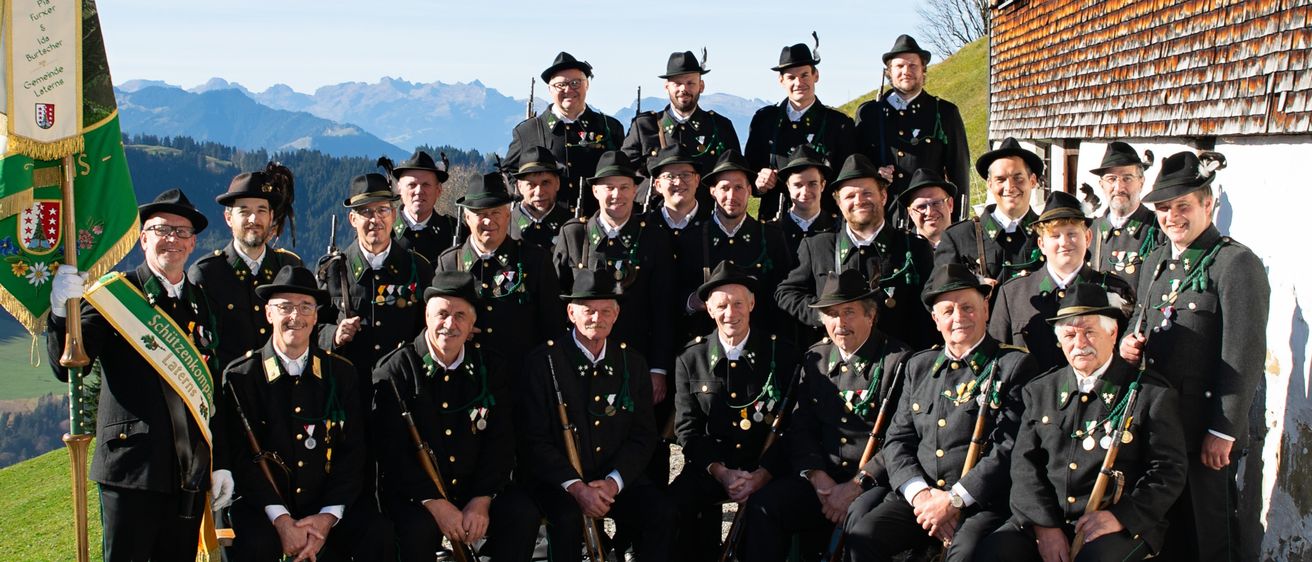 Eine Gruppe formell gekleideter Männer in Uniformen posiert für ein Foto vor einem Gebäude mit Bergen im Hintergrund.