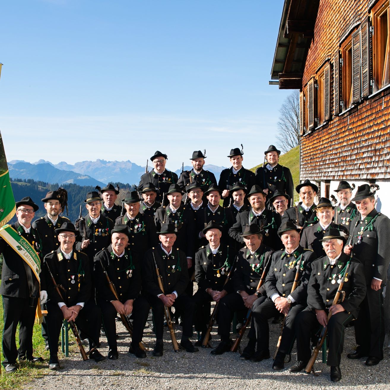 Eine Gruppe von Männern in Uniform, einige mit Gewehr, posiert für ein Foto mit Fahne und Gebäude im Hintergrund.