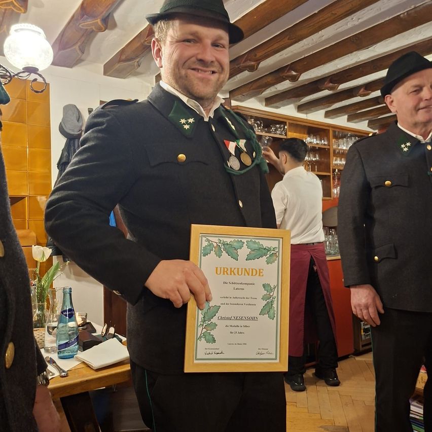 Ein Mann in Uniform hält ein gerahmtes Zertifikat im Innenbereich, während zwei weitere uniformierte Männer hinter ihm stehen. Im Vordergrund befindet sich ein Holztisch mit einer Blumenvase und einer Flasche.