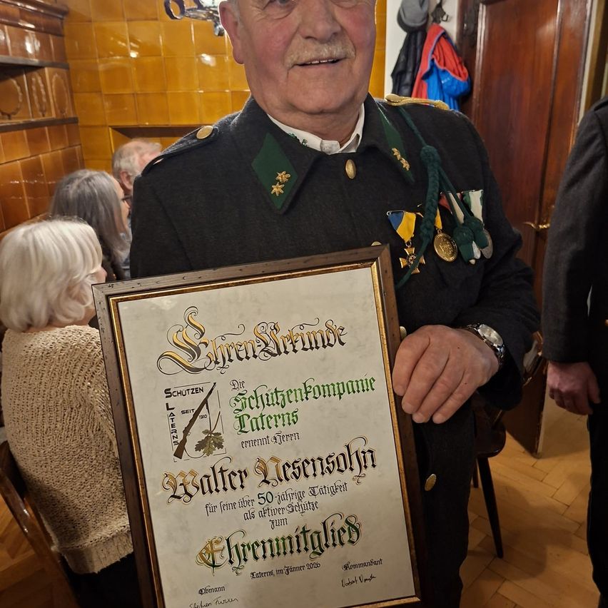 Ein Mann in Uniform hält ein Zertifikat, mehrere Leute stehen hinter ihm, einer mit Brille und einer mit weißen Haaren. Das Zertifikat ist gerahmt und hat goldene Ränder.