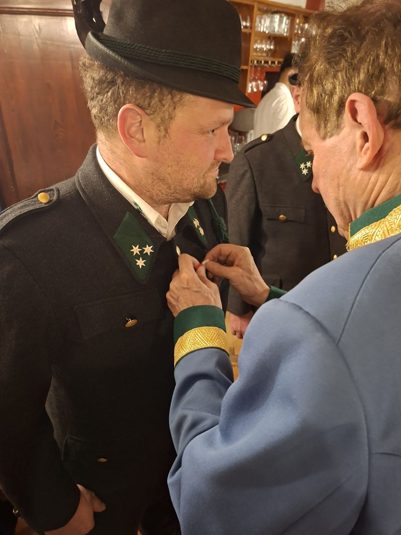 Ein Mann in Uniform bekommt von einem anderen Mann eine Medaille angesteckt. Sie befinden sich in einem Raum mit einer Holzwand.