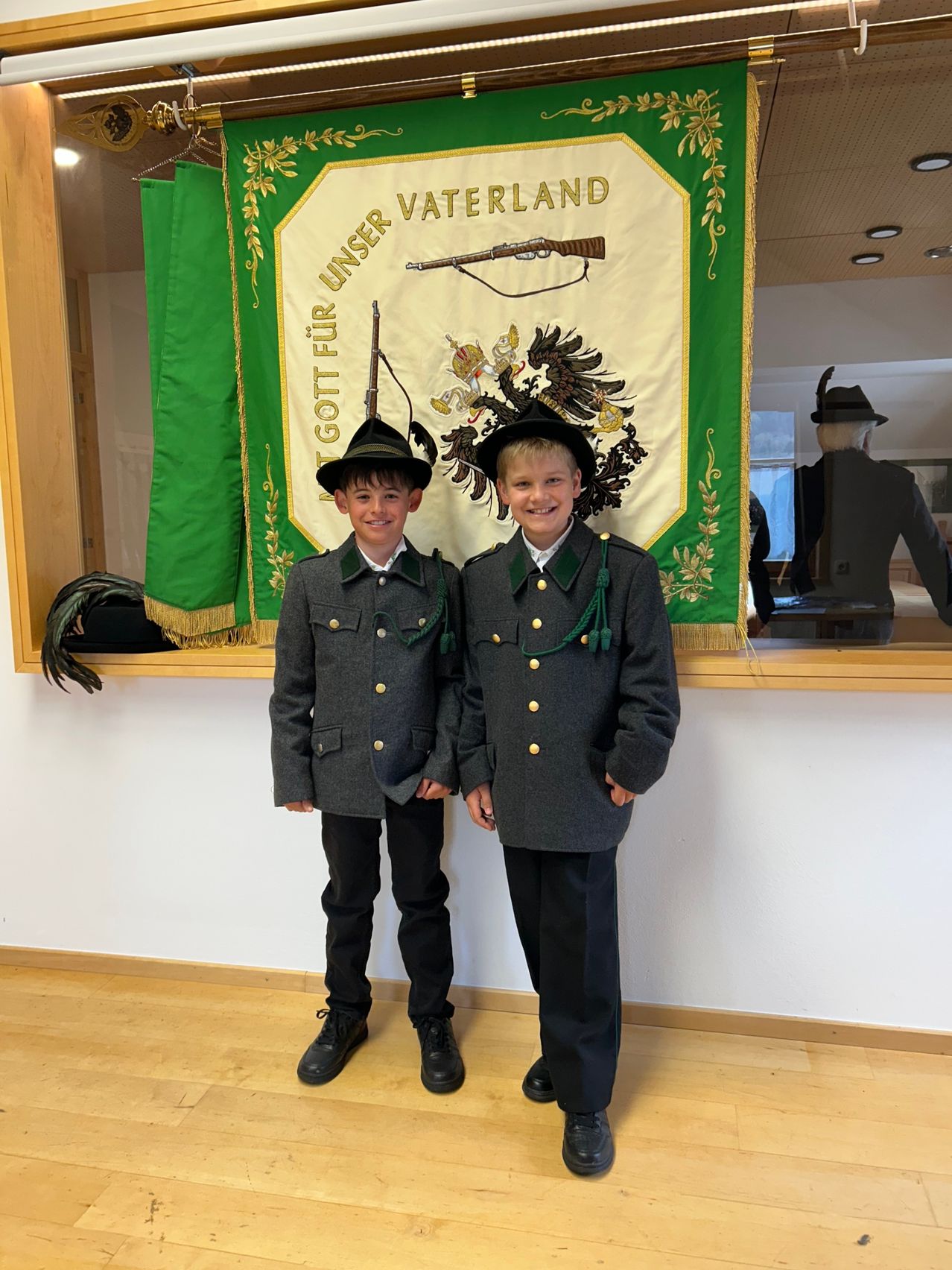 Zwei junge Jungen stehen zusammen vor einem Banner mit deutscher Schrift und einem Adlersymbol. Beide tragen Uniformen mit schwarzen Hüten und Schuhen.