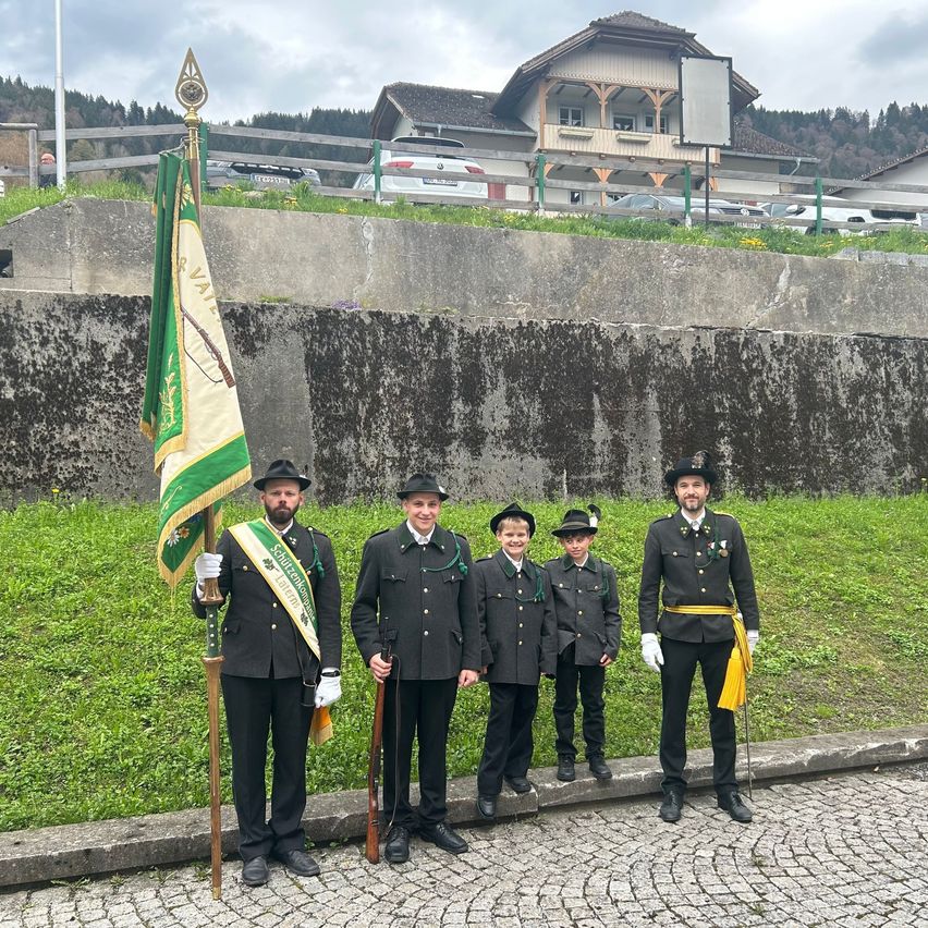 Fünf Männer in Uniform mit Gewehren stehen neben einer Flagge. Sie stehen auf einer Kopfsteinpflasterstraße mit einer Betonmauer und einem Grashügel hinter ihnen.