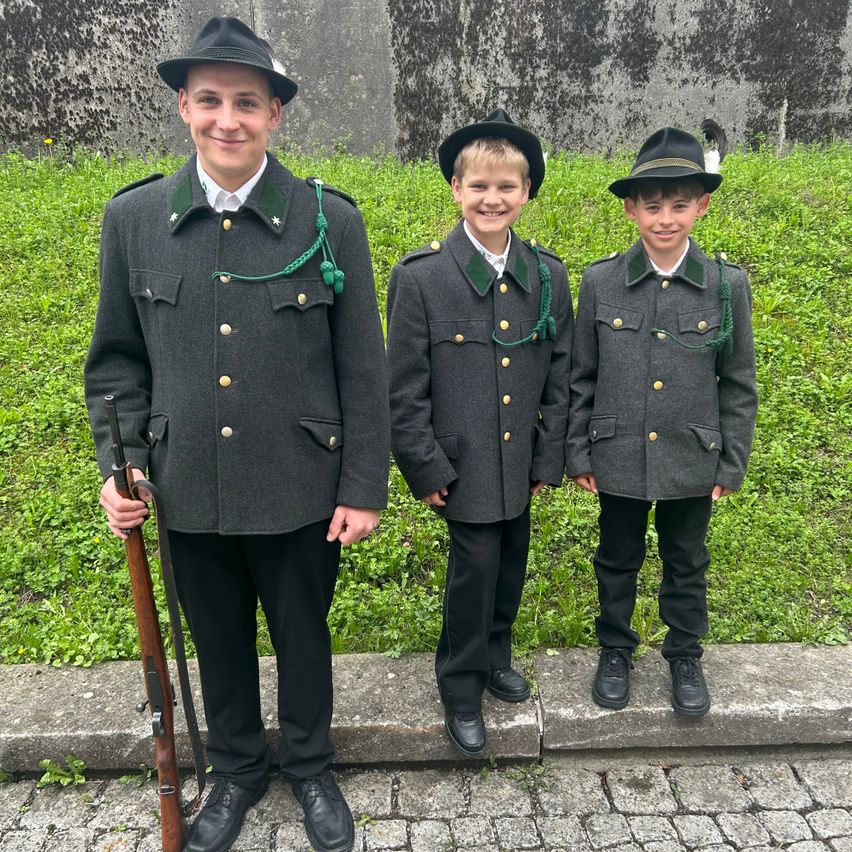 Drei junge Jungen in Uniformen stehen lächelnd zusammen auf einem Betonpfad. Der Junge auf der linken Seite hält eine Gewehr, und alle drei tragen Hüte und Stiefel. Dahinter befindet sich ein Rasenbereich und eine Steinmauer.