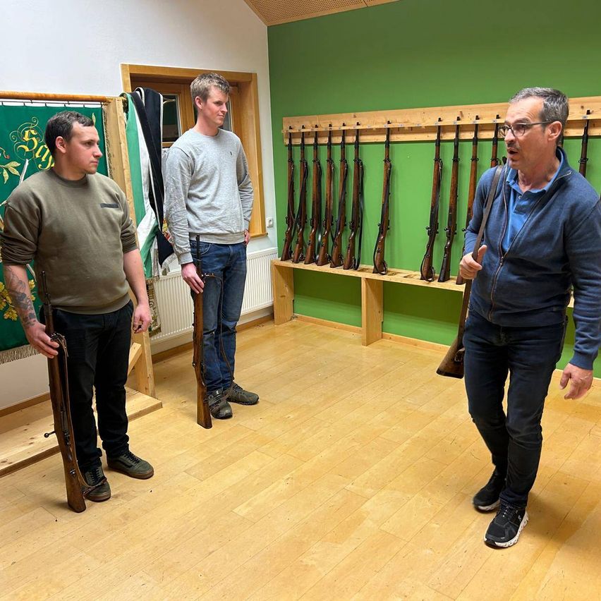 Drei Männer stehen in einem Raum mit grünen Wänden, jeder hält eine Waffe. Ein Mann spricht, während ein anderer zuhört. Gewehre sind an der Wand montiert.