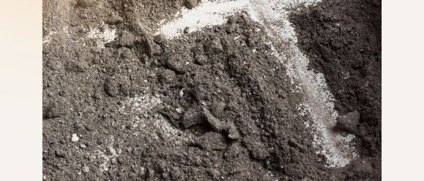 Eine Hand zeichnet mit einem Finger ein weißes Kreuz in die graue Asche. Der Satz Staub bist du und zum Staub kehrst du zurück ist im unteren Teil geschrieben.