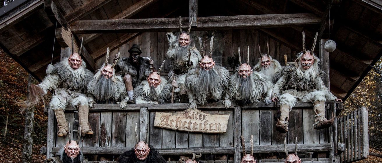 Eine Gruppe von Personen in aufwendigen, gehörnten Masken und Pelzkostümen posiert in einer rustikalen Holzscheune.
