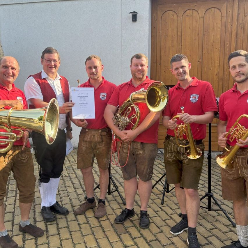 Eine Gruppe von Männern in traditioneller deutscher Kleidung, die Blechblasinstrumente halten, steht vor einer Holztür. Sie scheinen für ein Foto mit einem Zertifikat in der Mitte zu posieren.