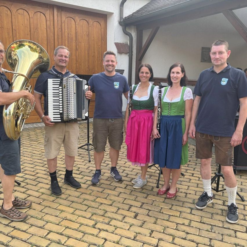 Eine Gruppe von Musikern steht vor einem Gebäude. Sie halten Instrumente wie Tuba, Akkordeon und Klarinette. Zwei Frauen tragen traditionelle Kleider, während die Männer lässig gekleidet sind.