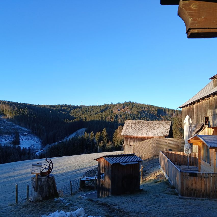 Eine frostige Landschaft mit einem Holzschuppen und einem kleinen Schild auf einem Baumstumpf, umgeben von schneebedeckten Bergen und Bäumen unter einem klaren blauen Himmel.