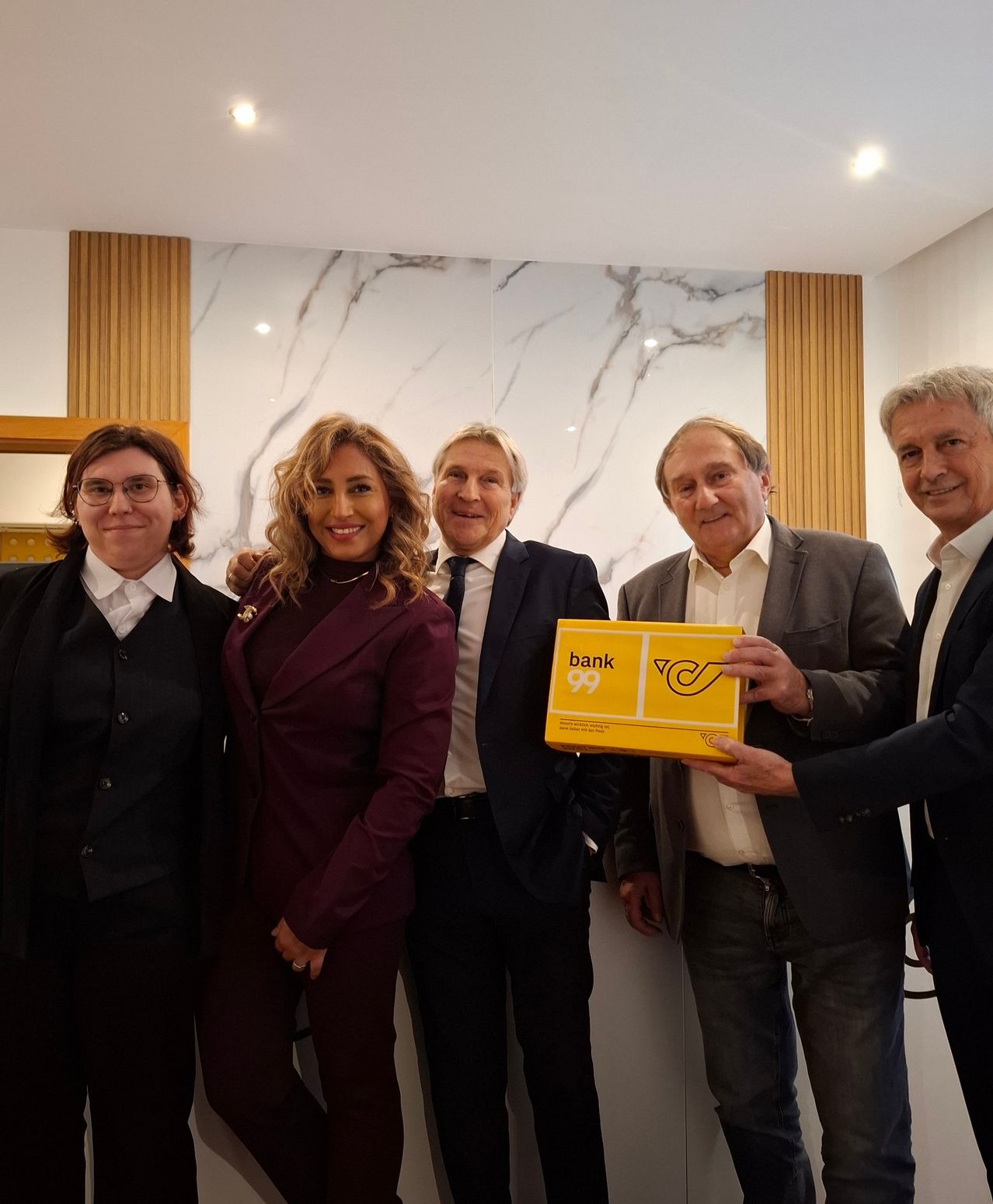 Fünf Personen stehen lächelnd vor einer Marmorwand. Die zentrale Person hält eine gelbe Box mit der Aufschrift 'Bank 99'. Zwei Männer tragen Anzüge und Krawatten, und zwei Frauen tragen Brillen.