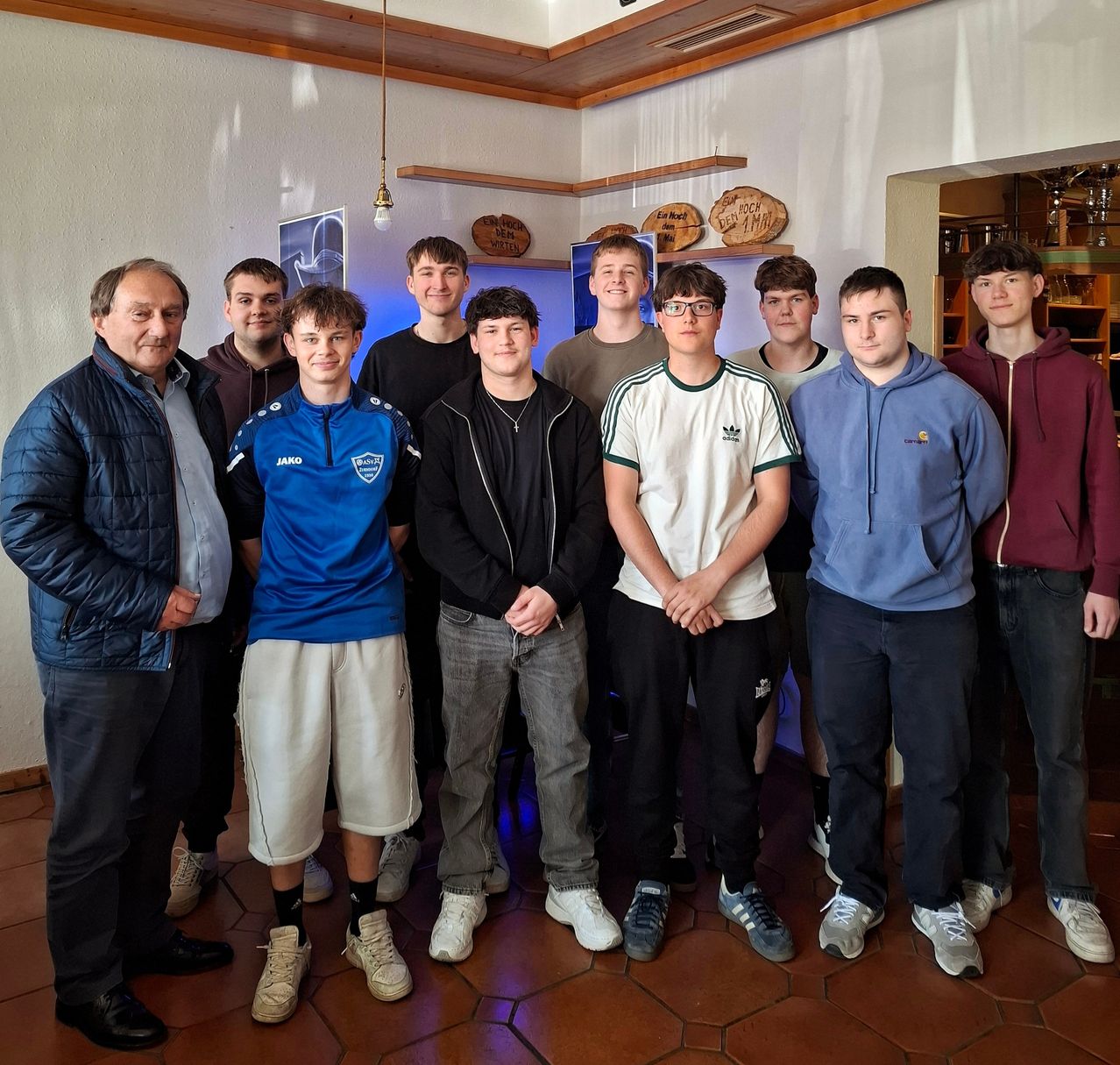 A group of young men and an older man pose for a photo in a room. The room has white walls and a tiled floor. Some of the men wear sneakers, and one wears glasses.