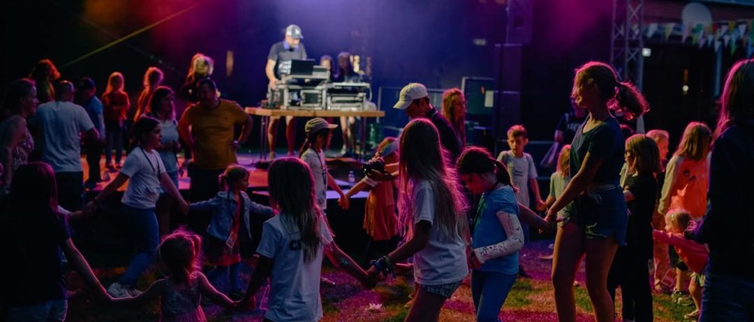 Eine Gruppe von Kindern und Erwachsenen tanzt auf einer Bühne mit bunten Lichtern. Einige Menschen stehen und schauen zu. Im Hintergrund spielt ein Mann Musik.