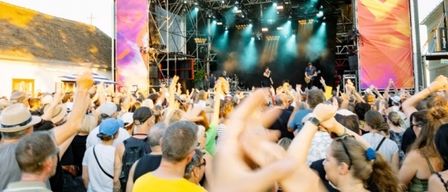 Ein lebhaftes Musikfestival mit einem tanzenden und jubelnden Publikum. Auf der Bühne spielen Musiker unter farbenfrohen Lichtern. Ein Hintergrund zeigt das Logo des Festivals.