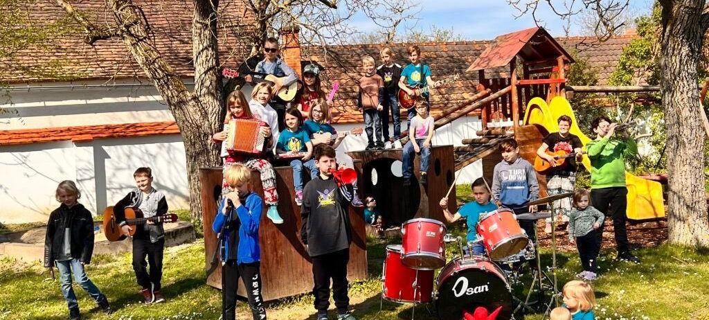 Eine Gruppe von Kindern und Jugendlichen ist draußen und einige spielen Instrumente. Gitarren, Trommeln und ein Akkordeon gehören zu den Instrumenten. Sie befinden sich auf einer Rasenfläche mit einem Baum und einem Spielhaus im Hintergrund.
