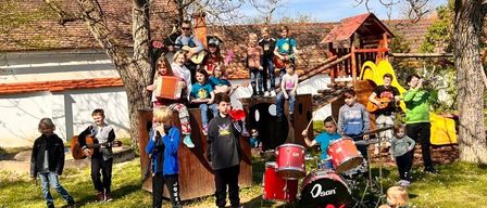 Eine Gruppe von Kindern und Jugendlichen ist draußen und einige spielen Instrumente. Gitarren, Trommeln und ein Akkordeon gehören zu den Instrumenten. Sie befinden sich auf einer Rasenfläche mit einem Baum und einem Spielhaus im Hintergrund.