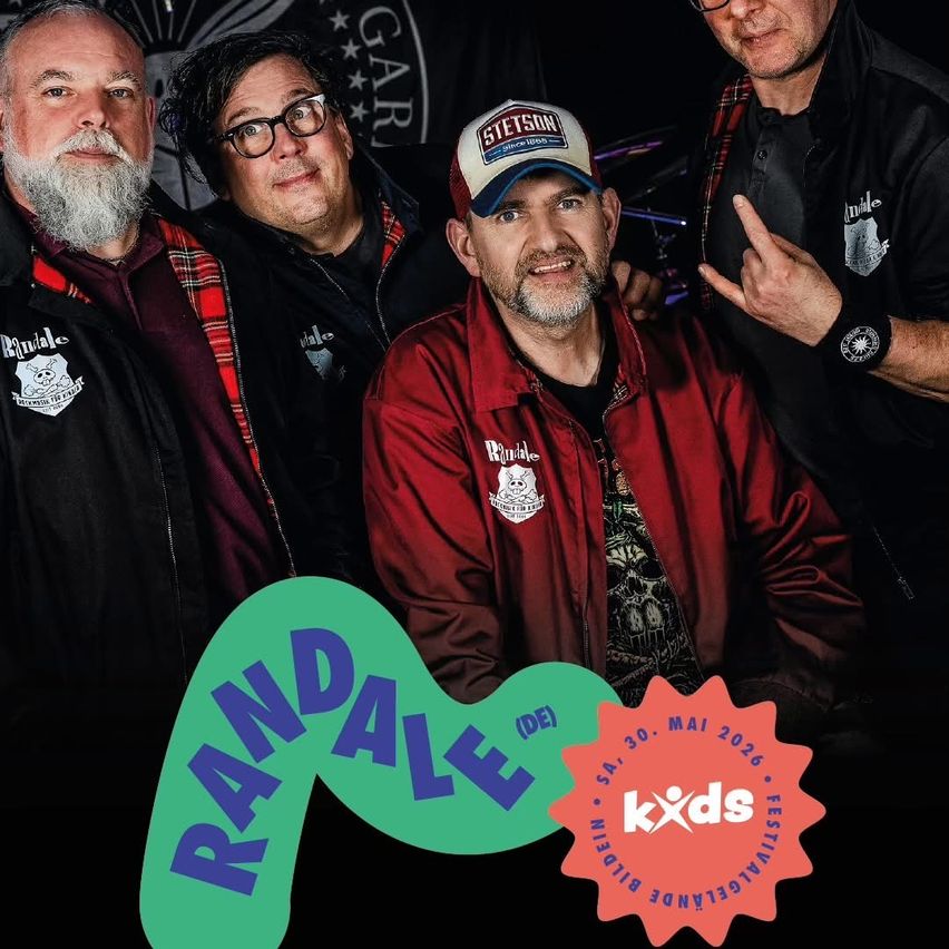 Vier Männer stehen zusammen für ein Foto. Der mittlere Mann trägt eine rote Jacke und eine Kappe mit dem Aufdruck 'STETSON'. Der Mann auf der linken Seite hat einen Bart und eine karierte Jacke. Der Mann auf der rechten Seite gibt ein Friedenszeichen. Im Hintergrund steht Text auf Deutsch.