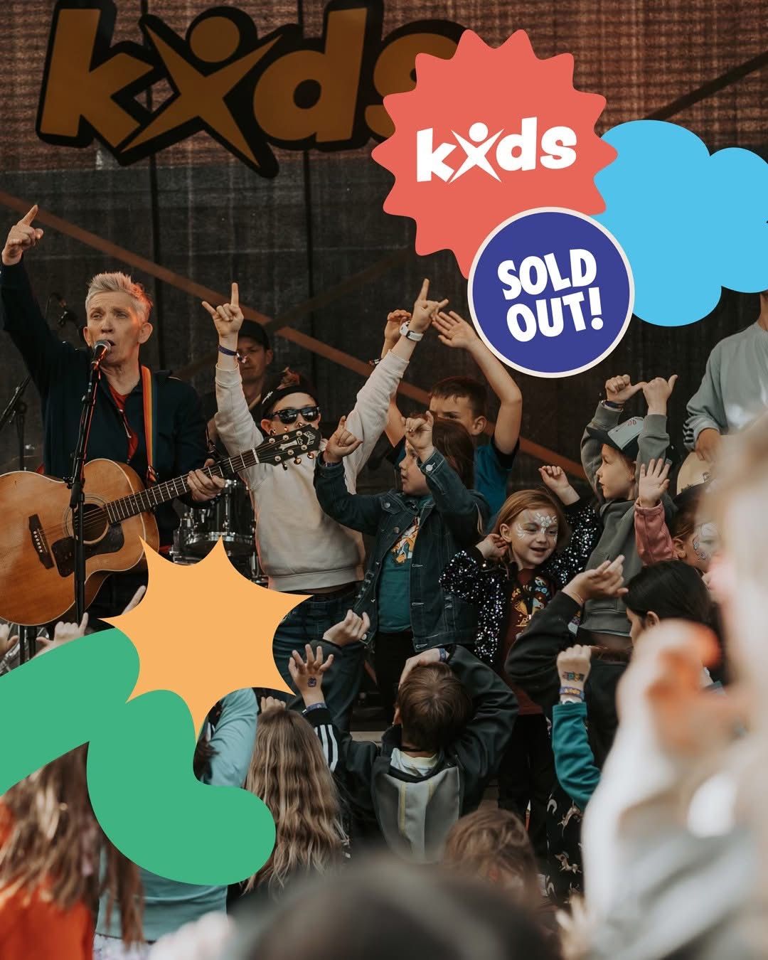 Eine Band spielt auf der Bühne vor einer Menge von Kindern und Erwachsenen. Der Leadsänger singt in ein Mikrofon, während eine andere Person Gitarre spielt. Das Publikum hebt die Hände und scheint die Musik zu genießen.