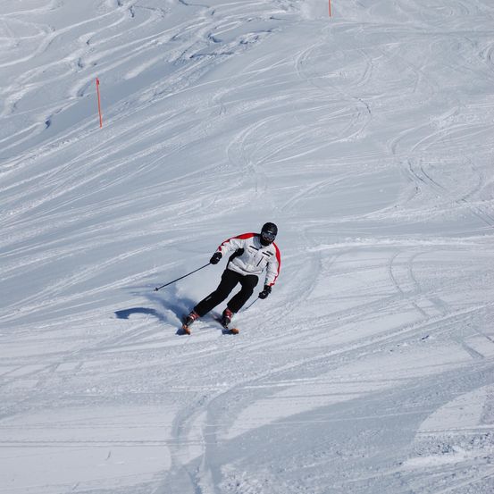 Ein Skifahrer in einem rot-weißen Anzug fährt einen schneebedeckten Hang hinunter und hält Skistöcke in der Hand.