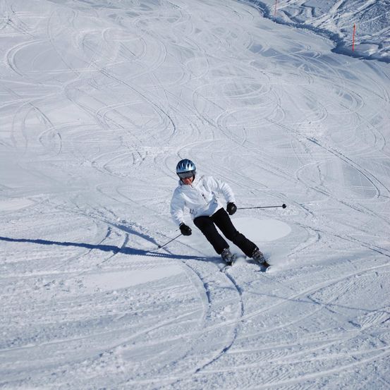 Ein Skifahrer, der eine weiße Jacke, schwarze Hosen und einen blauen Helm trägt, gleitet mit Skistöcken in der Hand den verschneiten Hang hinunter.