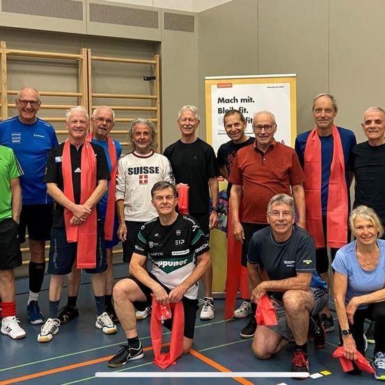 Eine Gruppe älterer Männer und Frauen posiert für ein Foto in einem Fitnessstudio. Ein Mann kniet und hält ein rotes Band. Das Banner hinter ihnen lautet 'Mach mit. Bleib fit.' Sie tragen Sportbekleidung.