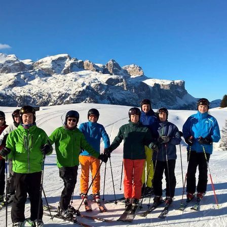 Eine Gruppe von Skifahrern posiert für ein Foto auf einem verschneiten Berg. Sie sind in Winterausrüstung und Helmen gekleidet, mit Bergen im Hintergrund, die in Schnee bedeckt sind.