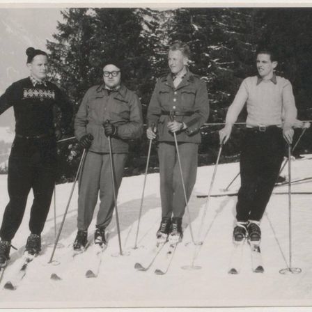 Vier Personen stehen auf einem verschneiten Hang und tragen Skiausrüstung. Sie lächeln für ein Foto. Im Hintergrund sind Kiefern und ein Berg zu sehen.