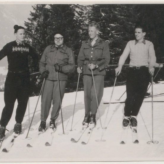 Vier Personen stehen auf einem verschneiten Hang und tragen Skiausrüstung. Sie lächeln für ein Foto. Im Hintergrund sind Kiefern und ein Berg zu sehen.