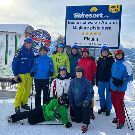 Gruppe von Skifahrern in bunter Winterkleidung posiert vor einem blauen Schild, das 'Skiresort.de' und 'Beste schwarze Abfahrt' besagt.