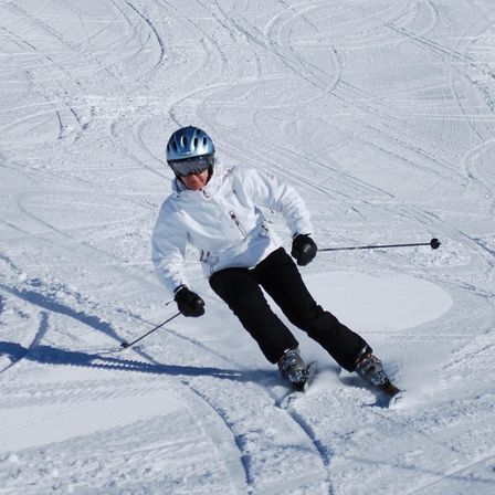 Ein Skifahrer in einer weißen Jacke und Helm fährt einen schneebedeckten Hang hinunter und hält Skistöcke.