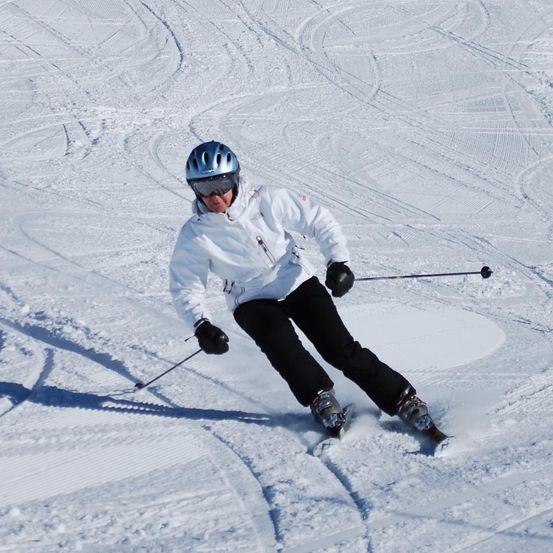 Ein Skifahrer in einer weißen Jacke und Helm fährt einen schneebedeckten Hang hinunter und hält Skistöcke.