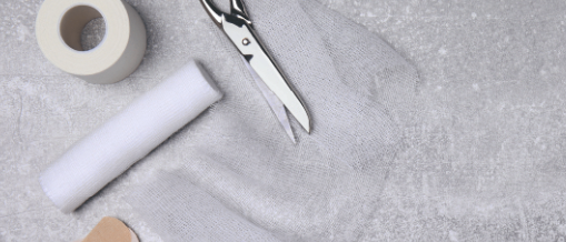 Ein Paar Scheren, eine Rolle Gaze und mehrere Verbände liegen auf einem grauen Hintergrund.