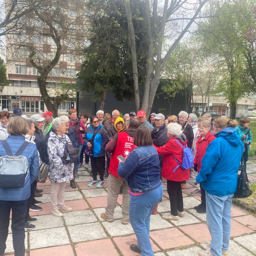 Eine Gruppe von Menschen versammelt sich in einem Park, einige tragen Hüte und haben Taschen dabei. Bäume und Gebäude sind im Hintergrund zu sehen.