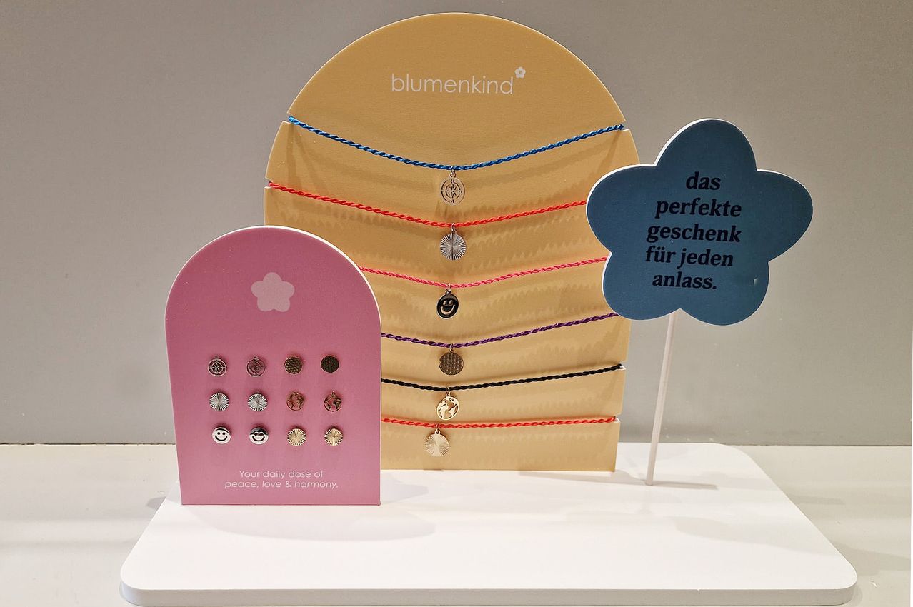 Eine Ausstellung von bunten blumenkind-Armbändern mit verschiedenen Anhängern auf einem Ständer, begleitet von einem Schild, das 'Das perfekte Geschenk für jeden Anlass' besagt.