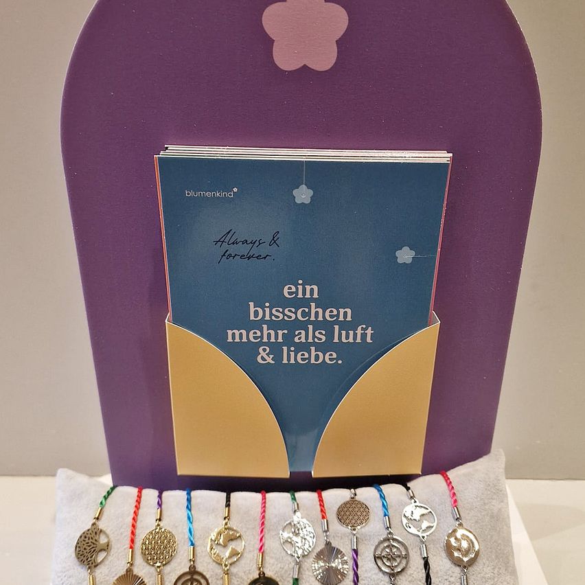 Ein violetter Rahmen hält ein Buch mit 'Always & Forever' darauf. Darunter ist ein goldener Umschlag mit dem deutschen Satz 'ein bisschen mehr als luft & liebe' geöffnet und zeigt bunte Armbänder darin.