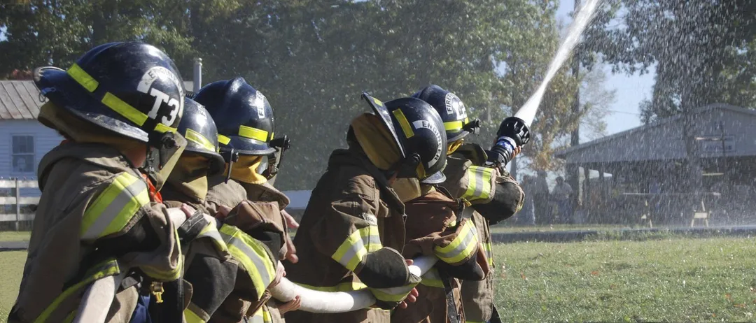 Eine Gruppe von Kindern in Feuerwehruniformen hält einen Schlauch und sprüht Wasser im Freien auf einem Rasen. Bäume und ein Gebäude sind im Hintergrund.