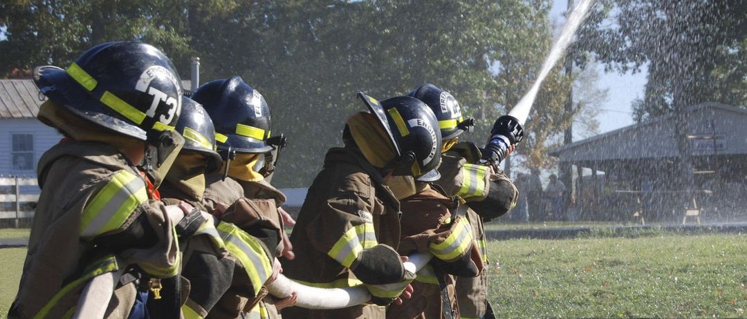 Eine Gruppe von Kindern in Feuerwehruniformen hält einen Schlauch und sprüht Wasser im Freien auf einem Rasen. Bäume und ein Gebäude sind im Hintergrund.
