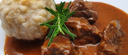 Ein Teller mit geschmortem Rindfleisch mit einer Beilage aus Kartoffelknödel und einem Zweig Rosmarin als Garnierung obenauf.