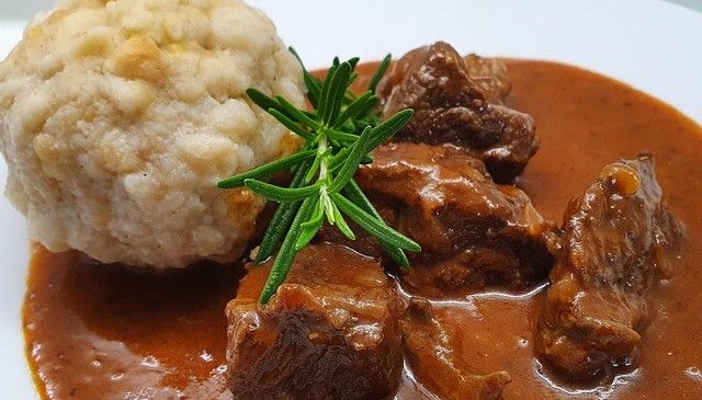 Ein Teller mit geschmortem Rindfleisch mit einer Beilage aus Kartoffelknödel und einem Zweig Rosmarin als Garnierung obenauf.