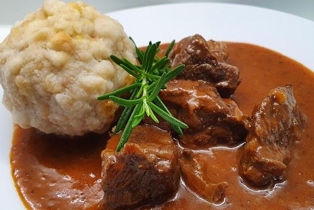 Ein Teller mit geschmortem Rindfleisch mit einer Beilage aus Kartoffelknödel und einem Zweig Rosmarin als Garnierung obenauf.