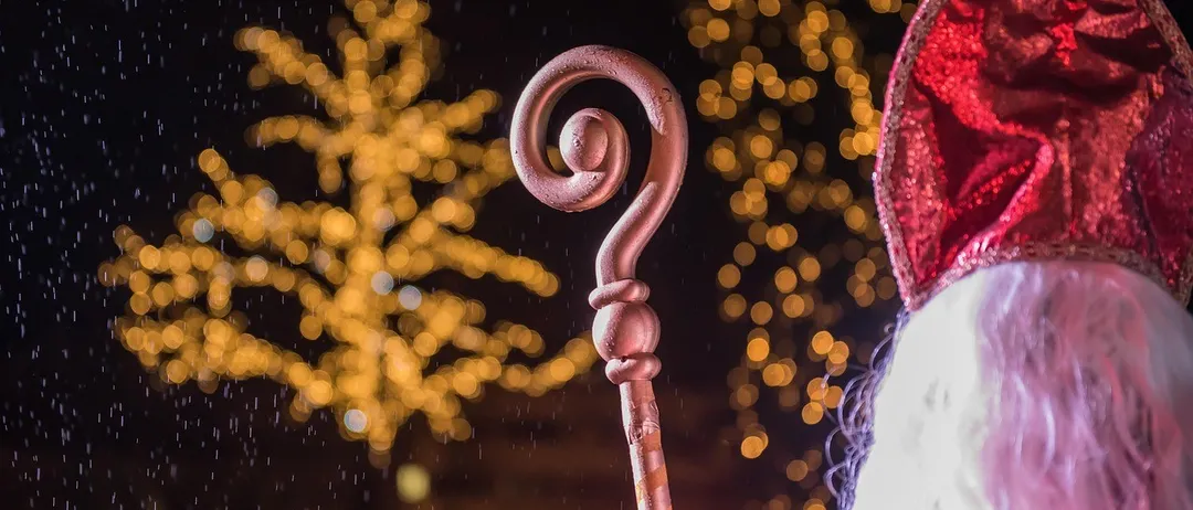 Eine verschwommene Aufnahme einer Person in Santa-Claus-Kleidung mit rotem Hut und rotem Umhang, die einen gebogenen Holzstab hält, und im Regen steht, mit beleuchteten Bäumen im Hintergrund.