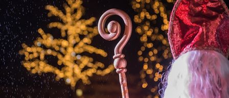 Eine verschwommene Aufnahme einer Person in Santa-Claus-Kleidung mit rotem Hut und rotem Umhang, die einen gebogenen Holzstab hält, und im Regen steht, mit beleuchteten Bäumen im Hintergrund.